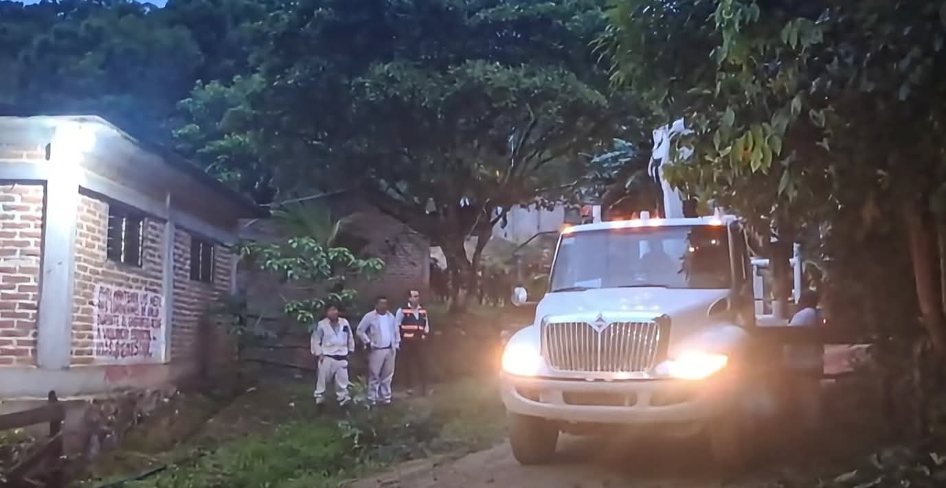 Camión blanco con grúa y trabajadores uniformados en un camino de terracería junto a una casa de ladrillo