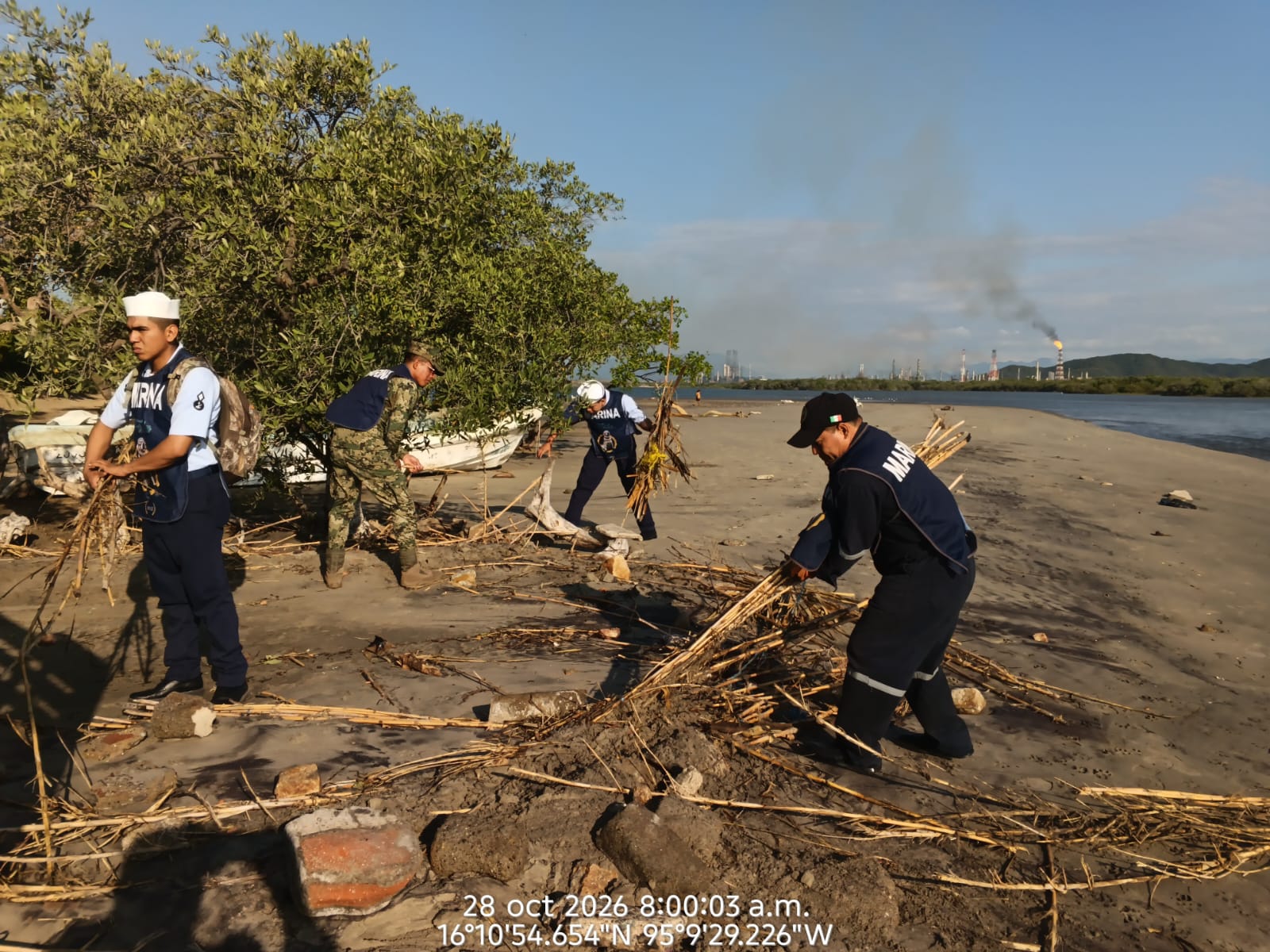 Elementos de Marina retiran ramas y desechos de la arena; al fondo se observa una refinería con una chimenea encendida