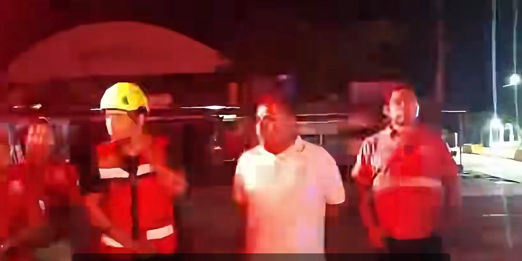 Personal con uniformes rojos y casco amarillo junto a un hombre de camisa blanca durante la noche