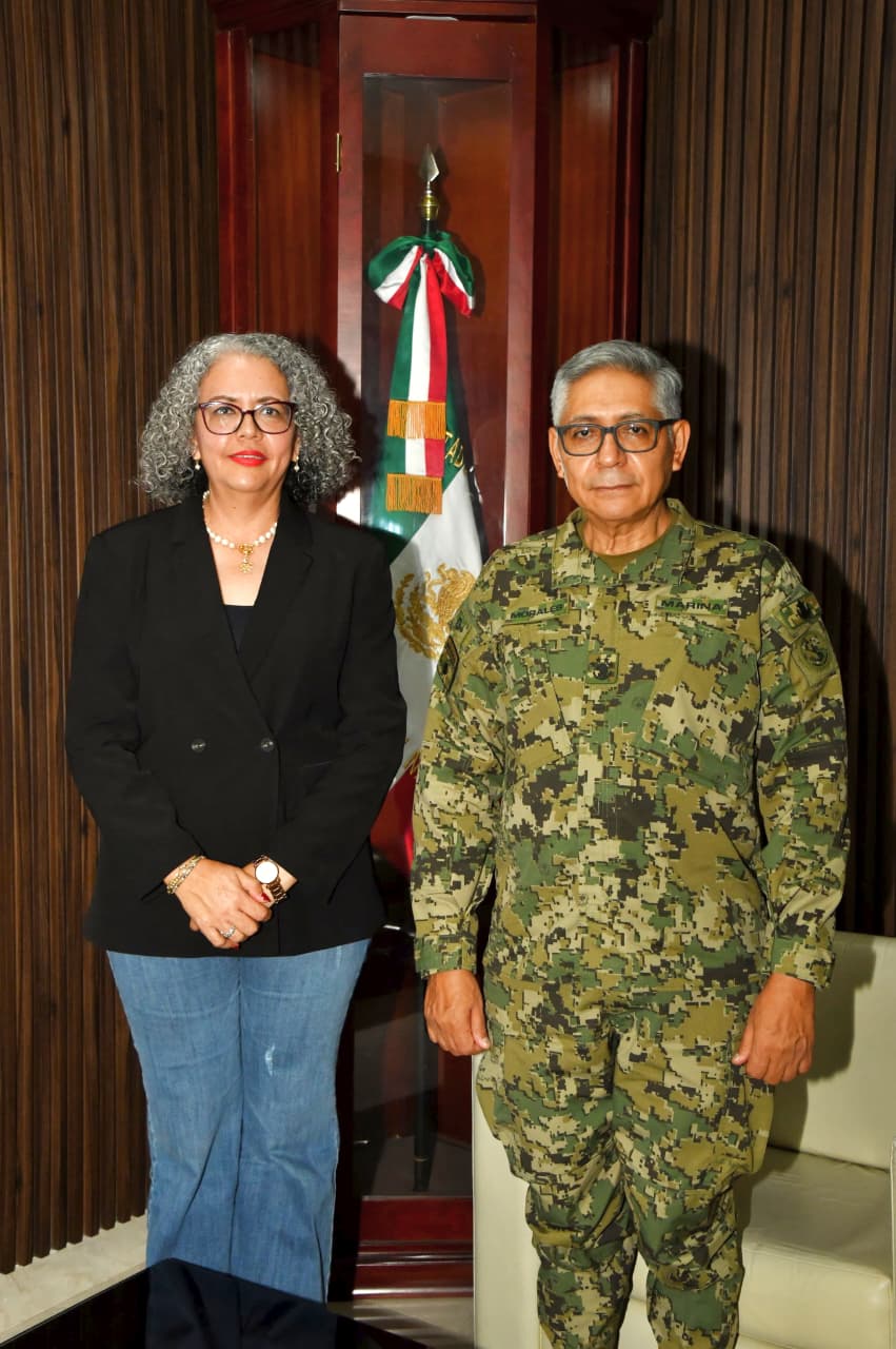 Un mando de la Marina con uniforme camuflado y una mujer de saco negro posan frente a una bandera de México en una oficina