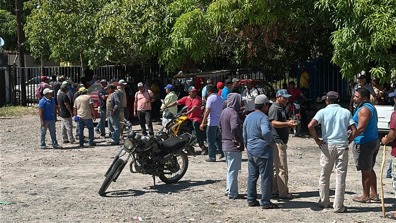 Campesinos reunidos a la sombra de árboles frente a una reja, con motocicletas estacionadas en el terreno