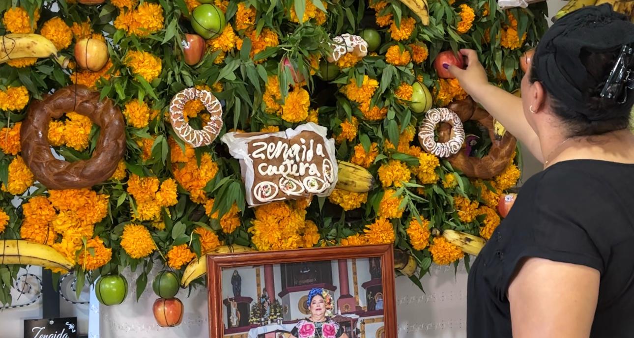 Mujer coloca una fruta en un altar cubierto de cempasúchil, panes de muerto y el retrato de una mujer con traje istmeño