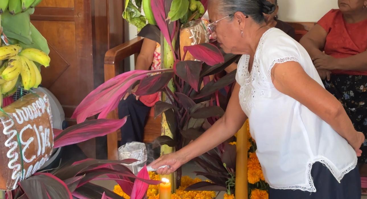 Mujer de blusa blanca enciende una vela frente a un altar con plátanos, hojas rojas y cempasúchil