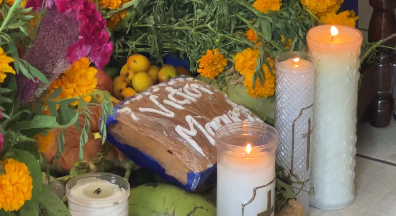 Pan de muerto con nombre escrito en betún, junto a veladoras encendidas, frutas y flores de cempasúchil