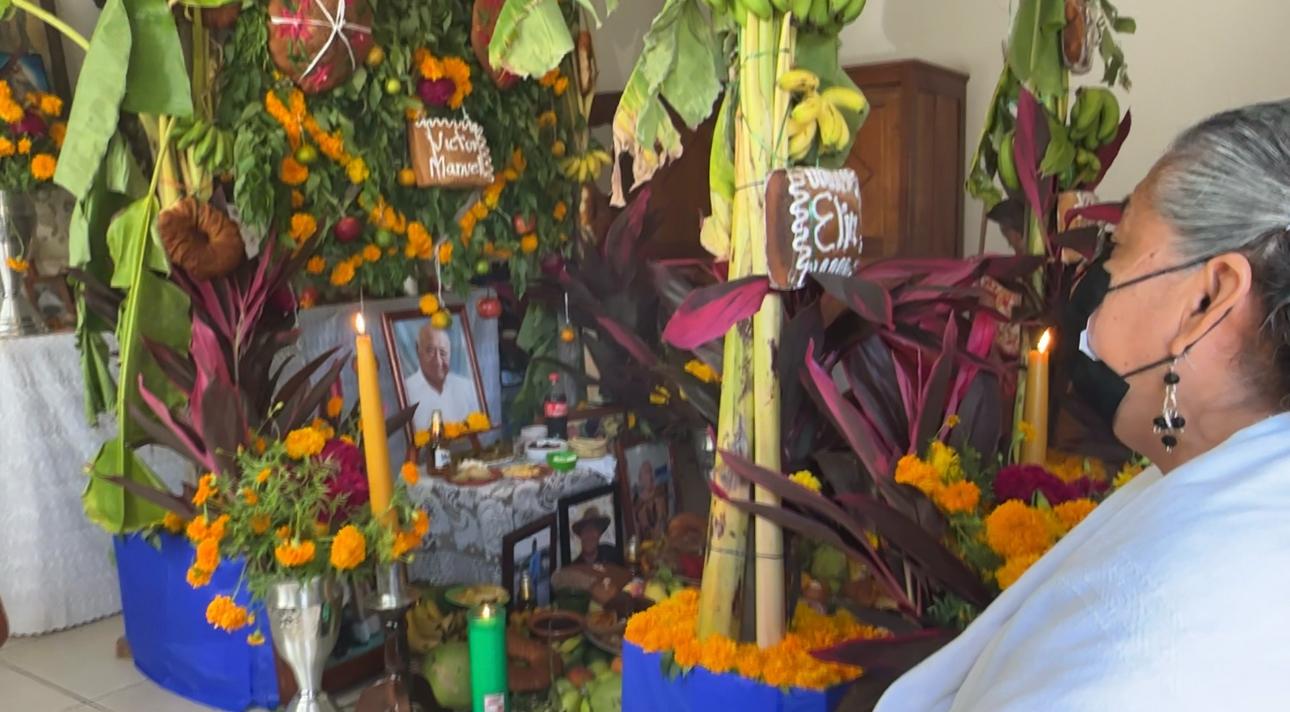 Mujer contempla un altar de muertos con retratos, velas, cempasúchil, cañas, plátanos y panes con nombres